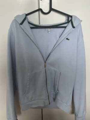 Ljusblå zip hoodie från Lacoste - Ljusblå hoodie från Lacoste med dragkedja framtill, klassisk huva och fickor på magen. Diskret Lacoste-logga på bröstet och gröna detaljer vid huvan. Tillverkad i mjukt bomullsmaterial för en skön och avslappnad look.