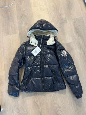 Mörkblå pufferjacka från Moncler - Snygg mörkblå pufferjacka från Moncler med glansig finish och avtagbar huva med ljus insida. Jackan har två dragkedjefickor fram och Moncler-logga på ärmen. Perfekt för kalla vinterdagar och riktigt trendig look.