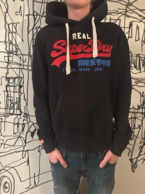 Superdry hoodie - Svart hoodie från Superdry med stor röd och blå logga på bröstet samt vita dragsnören i huvan. Klassisk känguruficka framtill och mjukt material som känns skönt mot huden. Perfekt för dig som gillar streetwear och vill ha en snygg och bekväm look.