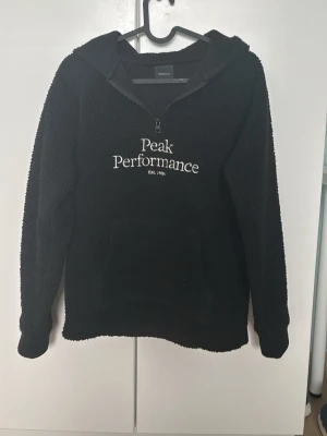 Svart fleecehoodie Peak Performance - Mysig svart fleecehoodie från Peak Performance med halv dragkedja och stor känguruficka framtill. Broderad logga i vitt på bröstet och huva för extra komfort. Perfekt för kyliga dagar och enkel att matcha med andra plagg.