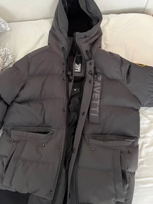 Mörkgrå dunjacka från Zavetti Canada - Snygg mörkgrå dunjacka från Zavetti Canada med stor huva, två stora framfickor med lock och tryckknappar samt patch på ärmen. Jackan har en hög krage, dragkedja och tryckknappar framtill samt tydlig logga på bröstet. Perfekt för kalla vinterdagar.