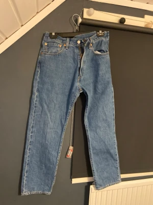 Levi's blå raka jeans - Klassiska blå Levi's jeans med rak passform och fem fickor. Jeansen har en hög midja, knappgylf och den ikoniska läderlappen bak i midjan. Tillverkade i slitstark denim som ger en tidlös look. Perfekta för dig som gillar vintage vibe och enkel stil. Storlek W32 L 30