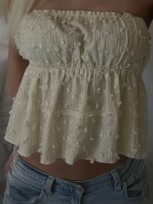 Ljusbeige ärmlös tubtopp med volang - Supersöt ljusbeige tubtopp med volang och små broderade prickar. Toppen är axelbandslös och har en luftig, flowy passform med resår upptill. Perfekt att styla med jeans eller kjol för en trendig look.
