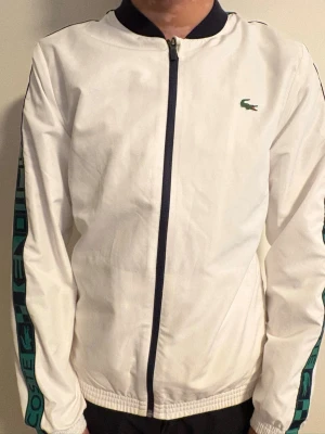 Vit träningsjacka från Lacoste Sport - Snygg vit träningsjacka från Lacoste Sport med marinblå dragkedja och detaljer. Jackan har ribbade muddar, ståkrage och grönt Lacoste-märke på bröstet. Ärmarna har gröna och blå ränder med Lacoste-logga. Perfekt för sport eller chill. Passar som XS också. Köparen står för frakten. Använt få gånger. Pris kan diskuteras. 