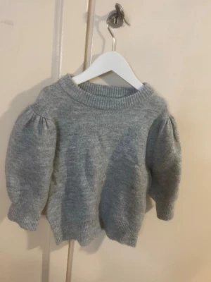 Grå stickad tröja med puffärm - Jag tycker den är 140-145. Mysig grå stickad tröja med bred ribbad halsringning och trendiga puffärmar. Tröjan har ribbade muddar vid ärmslut och nederkant, vilket ger en snygg och avslappnad look. Och enkel att matcha med alla slax byxor.