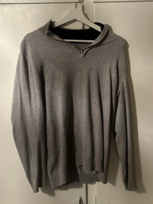 Grå hoodie med halv dragkedja - En enkel och stilren grå hoodie med halv dragkedja framtill och huva. Tillverkad i mjukt sweatshirtmaterial och har ribbade muddar vid ärmslut och nederkant. Perfekt för en avslappnad look.