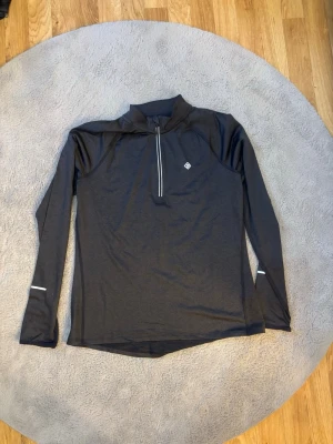 Mörkgrå långärmad träningströja - Snygg mörkgrå långärmad träningströja från Craft med halv dragkedja framtill och reflekterande detaljer på ärmar och rygg. Tillverkad i ett lätt och stretchigt funktionsmaterial som passar perfekt för löpning eller annan träning. Använd få tal gånger