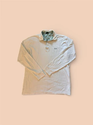 Vit långärmad skjorta från Hugo BOSS - Klassisk vit långärmad skjorta från Hugo BOSS med rutigt mönster på insidan av kragen och knappar framtill. Skjortan har en avslappnad passform och diskret BOSS-logga broderad på bröstet. Perfekt för en stilren och enkel look.