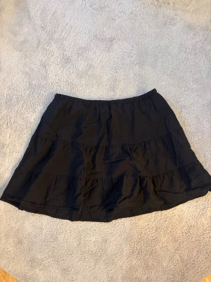 Svart volangkjol - Kort svart kjol med volangdetaljer och resår i midjan. Kjolen är gjord i mjuk bomull och har en enkel, flowy passform som ger en avslappnad och trendig look. Perfekt att matcha med en crop top eller hoodie. Använd få tal gånger