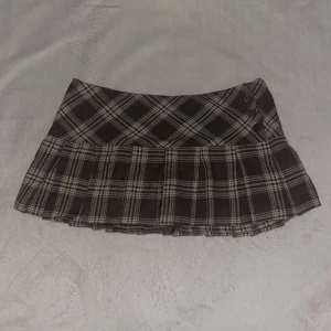 Brun rutig kortkjol med knappar - Snygg brun och beige rutig kortkjol med veckad nederdel och dekorativa knappar i midjan. Inbyggda shorts. Kjolen har en klassisk preppy stil och passar perfekt till både stickade tröjor och t-shirts.