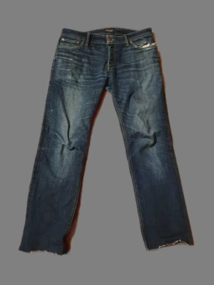 Jack&Jones  - Säljer ett par mörkblå jeans med rak passform och råa, avklippta benslut. Jeansen har klassiska fem fickor och slitna detaljer framtill. Perfekta för en avslappnad och trendig look.