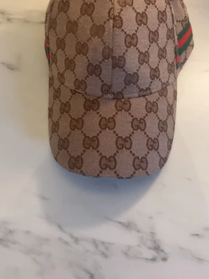 Gucci beige GG-mönstrad keps - Säljer en beige keps från Gucci med det klassiska GG-monogrammet över hela kepsen. Kepsen har justerbart spänne bak i metall med Gucci-logga och en grön-röd rand på sidan. Materialet är canvasliknande och kepsen har böjd skärm.