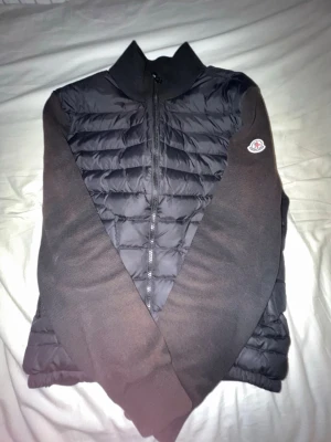 Svart cardiganjacka från Moncler - Snygg svart cardiganjacka från Moncler med quiltad framsida och stickade ärmar. Jackan har dragkedja framtill, ribbad krage och Moncler-logga på ena ärmen. Perfekt för lager-på-lager och stilren look. Lite solblekt också men går forfarande att använda! Priset kan diskuteras