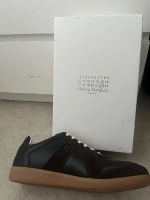 Maison Margiela svarta sneakers - Maison Margiela sneakers i svart och mörkbrunt med ovandel i mocka och läder, vita skosnören och klassisk gummisula. Snygg och stilren design som passar till det mesta. Kommer med originalkartong.