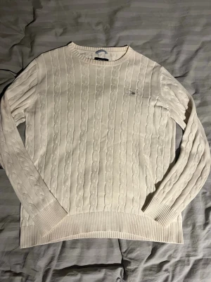 Vit gant tröja - Kabelstickad vit tröja från Gant med rund halsringning och diskret logga på bröstet. Tröjan har långa ärmar och ribbade muddar vid ärmslut och nederkant. Klassisk och stilren design i mjukt material.