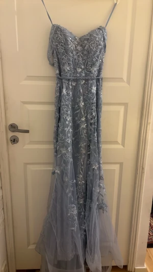 Ljusblå långklänning med spetsdetaljer - Elegant ljusblå långklänning med tunna axelband och vackra spetsdetaljer i blommönster. Klänningen har en figurnära passform upptill och faller mjukt nedåt med ett skirt, lager på lager-tyg. Perfekt för bal eller festliga tillfällen.