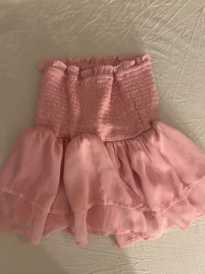 Rosa volangig bandeau tubtopp - Supersöt rosa bandeau tubtopp med smockad överdel och dubbla volanger nedtill. Toppen är ärmlös och har en romantisk vibe, perfekt för sommarens alla tillfällen. Tillverkad i ett lätt och luftigt material som känns skönt mot huden.