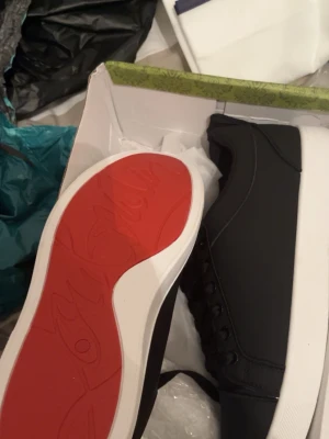 Svarta sneakers med röd sula - Snygga svarta sneakers från Christian Louboutin med klassisk röd sula och vita kanter. Skorna har en stilren design med svarta snören och diskreta sömmar. Perfekta för dig som vill ha något trendigt och exklusivt. Lite smutsiga bara vid sulan men kan fixa rent 