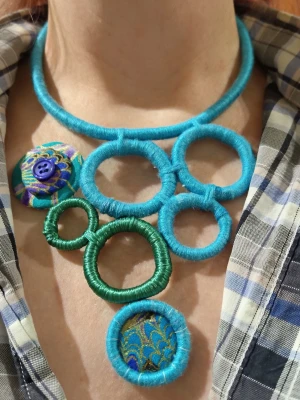 Färgstarkt halsband med garnklädds ringar - Unikt halsband med stora ringar klädda i blått och grönt garn. En ring har mönstrat tyg och en dekorativ knapp. Halsbandet har en lekfull och kreativ design med olika former och färger, perfekt för dig som gillar att sticka ut.
