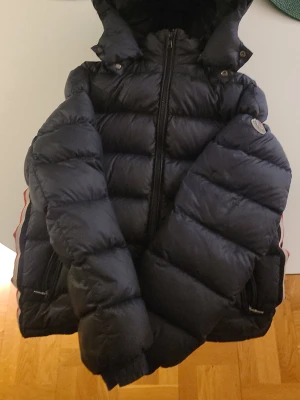 Svart pufferjacka från Moncler - Snygg svart pufferjacka från Moncler med huva och dragkedja framtill. Jackan har Moncler-logga på ärmen och coola röda och vita detaljer längs sidorna. Fylld med dun för extra värme och har två fickor med dragkedja. Perfekt för kalla vinterdagar.