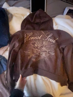 Brun zip up hoodie Honest Soul med tryck - Säljer en brun hoodie med dragkedja från Divided. På ryggen finns ett stort, stilrent tryck med texten 'Honest Soul' och 'Love' i ett band, samt grafiska detaljer. Hoodien har huva och långa ärmar, perfekt för en chill och avslappnad look. Skit snygg!
