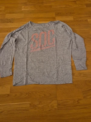 Grå långärmad tröja från SOC - En grå långärmad topp från SOC med stort rosa tryck på bröstet. Tröjan har en avslappnad passform, bred halsringning och är tillverkad i ett mjukt, tunt material som känns skönt mot huden. Perfekt för en chill och sportig look.