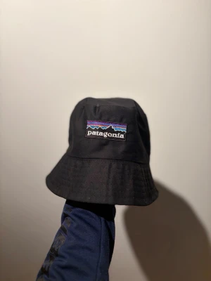 Svart bucket hat från Patagonia - Snygg svart bucket hat från Patagonia med klassisk broderad logga framtill i vitt, blått och lila. Hatten har mjuk form och bred skärm som ger en avslappnad look. Tillverkad i slitstarkt tyg, perfekt för att lyfta din outfit.