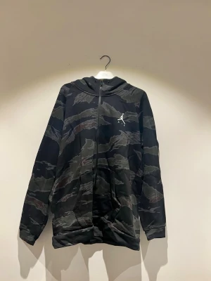 Jordan camo zip hoodie med huva - Snygg hoodie från Jordan med hel dragkedja och huva. Camouflage-mönster. Ikoniska Jumpman-loggan i vitt på bröstet. Mjukt material och två fickor framtill. Perfekt för en streetwear-look.