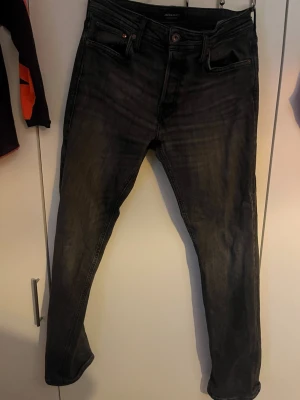 Svarta slim fit jeans från Jack & Jones - Säljer ett par svarta slim fit jeans från Jack & Jones med klassisk femficksdesign och snygga slitningar. Jeansen har en knappgylf och är tillverkade i bomull med lite stretch för skön passform. Perfekta för dig som gillar en modern och stilren look.