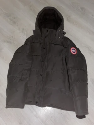 Svart dunjacka från Canada Goose - Säljer en svart dunjacka från Canada Goose med klassisk patch på ärmen. Jackan har huva, dragkedja och knappar framtill samt en stor ficka på bröstet. Perfekt för kalla vinterdagar och riktigt varm tack vare dunfyllningen.