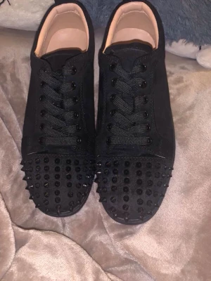 Svarta Louboutin sneakers med nitar - Säljer ett par svarta sneakers från Christian Louboutin med coola svarta nitar på tån och den ikoniska röda sulan. Skorna har svarta skosnören och en stilren design i mocka och läder. Perfekta för dig som vill sticka ut med din stil.