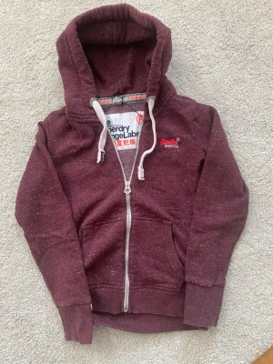 Vinröd hoodie från Superdry - Snygg vinröd hoodie från Superdry med vitmelerad effekt och broderad logga på bröstet. Tröjan har huva med vita snören, hel dragkedja och praktiska fickor framtill. Perfekt för dig som gillar en avslappnad och cool stil.