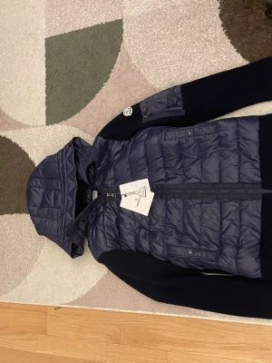Mörkblå dunjacka från Moncler - Snygg mörkblå dunjacka från Moncler med huva och stickade ärmar. Jackan har dragkedja framtill, två fickor med knapp och en Moncler-logga på ärmen. Perfekt för våren