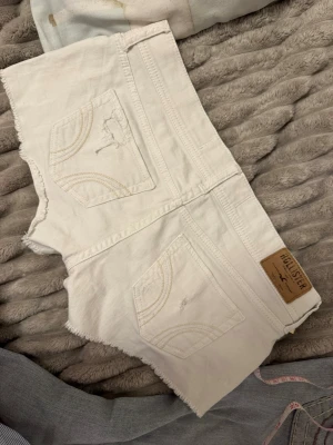 Vita jeansshorts från Hollister - Säljer ett par vita jeansshorts från Hollister med råa kanter och slitna detaljer. Shortsen har klassiska bakfickor med broderade sömmar och en liten logga fram. Perfekta för sommaren och har en låg midja för en avslappnad look.