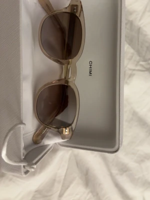 Beige solglasögon från CHIMI - Snygga solglasögon från CHIMI med transparent beige båge och bruna tonade glas. Klassisk rundad form som passar till många stilar. Kommer med originalfodral och putsduk med CHIMI-logga. Perfekta för soliga dagar och trendiga outfits.