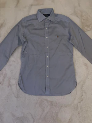 Polo Ralph Lauren skjorta - Klassisk ljusblå skjorta från Polo Ralph Lauren med knappstängning framtill och broderad logga på bröstet. Skjortan har lång ärm och normal passform, perfekt till en stilren look. Tillverkad i bomull för en bekväm känsla.