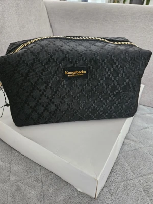 Svart necessär från Kungsbacka - Snygg svart necessär från Kungsbacka med diskret rutmönster och guldfärgad dragkedja. Tillverkad i slitstarkt tygmaterial och har en rektangulär form som rymmer allt du behöver till resan eller vardagen. Perfekt för att organisera dina småprylar.