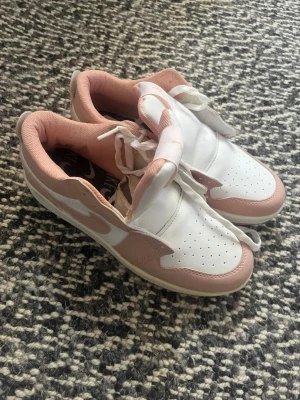 Jordan sneakers i vitt och rosa - Snygga sneakers från Jordan i vitt och ljusrosa med klassisk logga på plösen. Skorna har lågt skaft, perforerad tå och vita skosnören. Ovandelen är i läder och mesh för en fräsch look. Perfekta för dig som vill ha en clean och sportig stil.