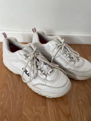 Vita Fila Disruptor sneakers - Chunky vita Fila Disruptor sneakers med klassisk retrostil. Skorna har tjock sula, snörning och detaljer i rött och blått på sidan och plösen. Tillverkade i syntetmaterial med textilfoder och dragflik bak. Perfekta för dig som gillar streetwear och sportig look. De har synbara slitage på visa delar av skon. Som man kan se på bilderna men har ändå mer att ge. 