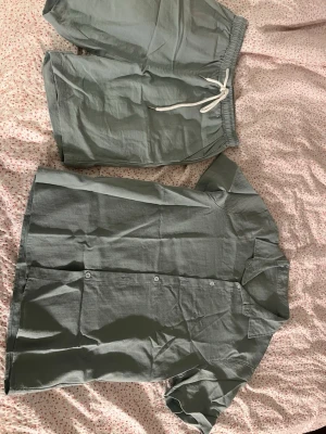 Sett - Säljer ett stilrent grönt linneset med kortärmad skjorta och matchande shorts. Skjortan har knappar framtill och klassisk krage, shortsen har resårmidja med vit dragsko. Perfekt för chill hemma eller mysiga kvällar.