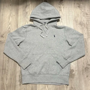 Ralph Lauren hoodie  - Snygg grå hoodie från Polo Ralph Lauren med klassisk broderad logga på bröstet. Hoodien har huva med vita dragsnören och en stor känguruficka framtill. Tillverkad i mjukt bomullsmaterial och har ribbade muddar vid ärmslut och nederkant.