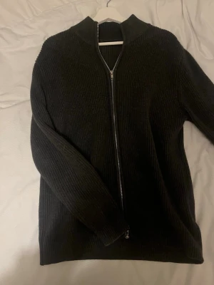 Mörkgrå ribbstickad ziptröja - Säljer en mörkgrå ribbstickad tröja med dragkedja framtill och hög krage. Tröjan har långa ärmar och är gjord i ett mjukt, tjockare material som passar perfekt när det är kyligt ute. Enkel och stilren design som är lätt att matcha.