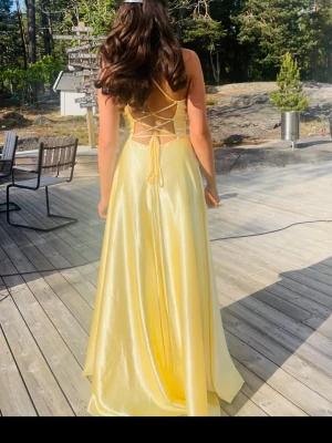 Ljusgul balklänning 💛 - Elegant ljusgul långklänning i glansigt material med smala axelband och v-ringad front. Klänningen har öppen rygg med snygg snörningsdetalj. Endast andvänd en gång, kan behöva tvättas då det kan vara lite smutsigt nertill💕Pris kan dissekeras!