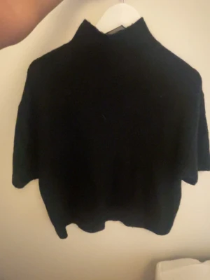 Svart stickad kortärmad tröja - En svart stickad tröja med korta ärmar och hög hals. Tröjan har en boxig och avslappnad passform, perfekt för lager-på-lager. Materialet ser mjukt och mysigt ut, vilket gör den skön att bära under kyligare dagar.