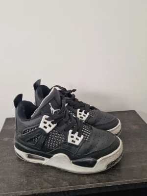 Svarta Air Jordan 4 Retro sneakers - Snygga svarta Air Jordan 4 Retro sneakers med vita och svarta detaljer. Skorna har klassisk meshpanel på sidorna, perforerad tå och ikoniska plastdetaljer vid snörningen. Tjock sula med synlig Air-enhet och Jumpman-logga på plösen.