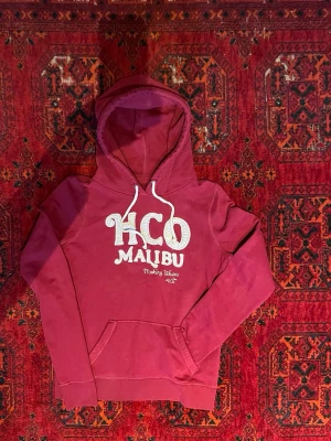 Hollister Hoodie - Säljer en röd hoodie från HCO med stor vit text 'HCO MALIBU' och broderad detalj 'Making Waves' framtill. Hoodien har klassisk känguruficka och vita snören i huvan. Perfekt för dig som gillar avslappnad och cool stil. Undrar du något är det bara att skriva😁🙌
