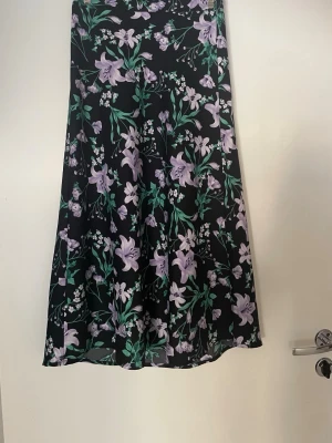 Svart midikjol med lila blommor - Snygg svart midikjol i stl 34 med  lila och grönt blommönster. Kjolen har en rak och flowy passform som ger en avslappnad men ändå stilren look. Perfekt att matcha med en enkel topp för en trendig outfit. Lila linne från Vila i stl S. Säljes tillsammans.