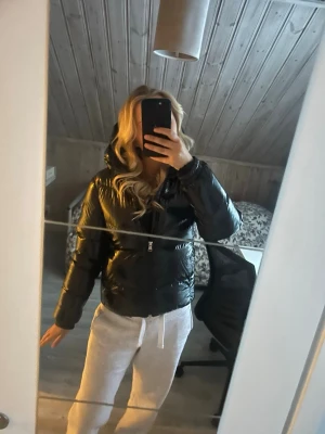 Svart glansig pufferjacka med huva - Säljer en svart, glansig pufferjacka med dragkedja och huva. Jackan är kortare i modellen och har en modern, boxig passform. Perfekt för kalla dagar när du vill hålla stilen och värmen.