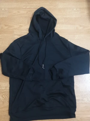 Svart basic hoodie med huva - En enkel svart hoodie med huva och dragsko. Hoodien har en stor magficka framtill och ribbade muddar vid ärmslut och nederkant. Perfekt för en avslappnad och stilren look. Materialet känns mjukt och bekvämt, troligen bomullsblandning.