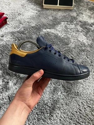 Adidas Stan Smith X Raf Simons sneakers - Mörkblå Adidas Stan Smith sneakers i läder med gul detalj på hälen och matchande blå skosnören. Klassisk låg modell med perforerade ränder på sidan och svart sula. Perfekta för dig som gillar stilrena och sportiga sneakers.